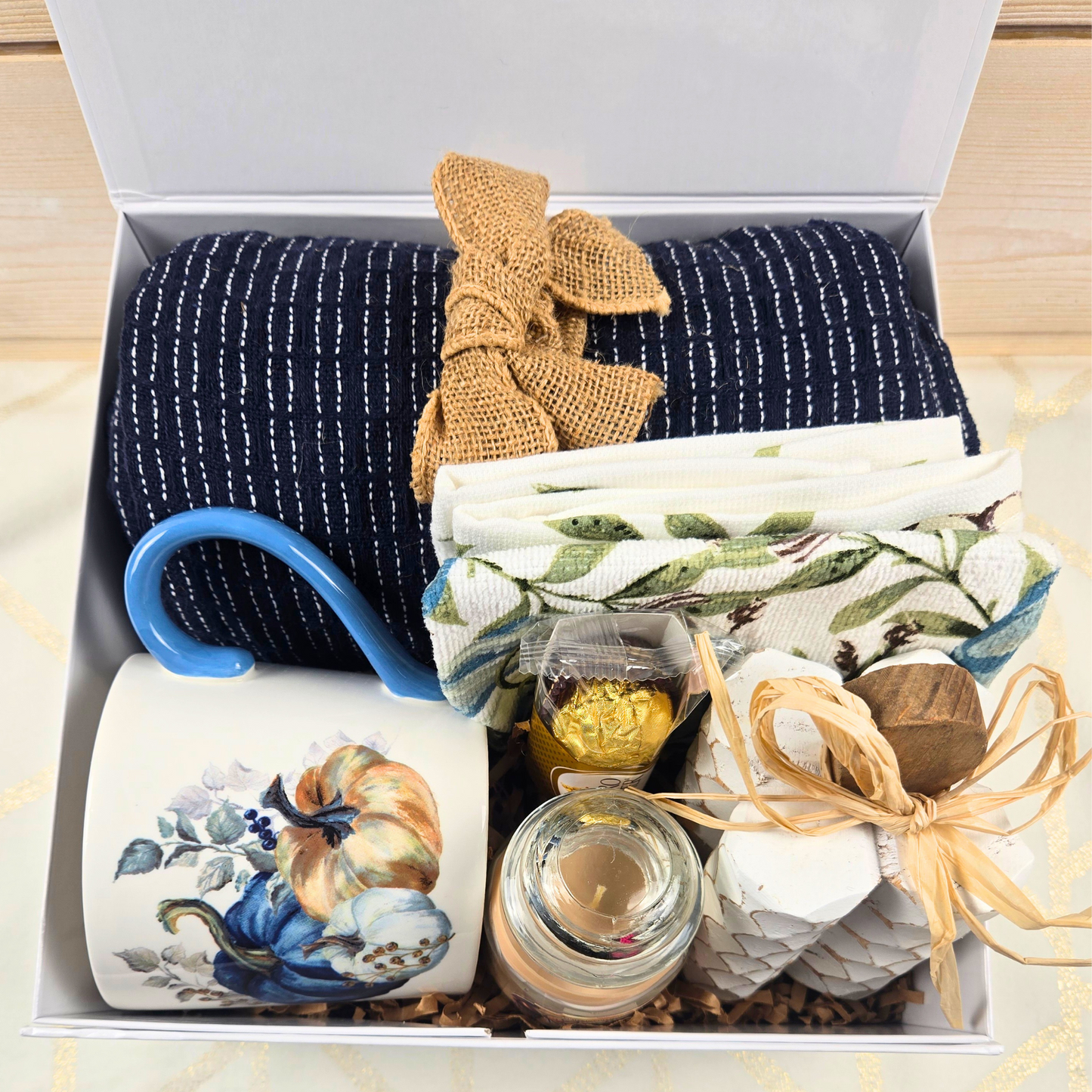 Cozy Fall Gift Box for Her with Elegant Blue Accents and Magnet Closure with navy blue throw, 16 oz mug, blue pumpkin dish towel, 3 oz pumpkin maple custard candle, white pumpkin decor and Ferrero Rocher.