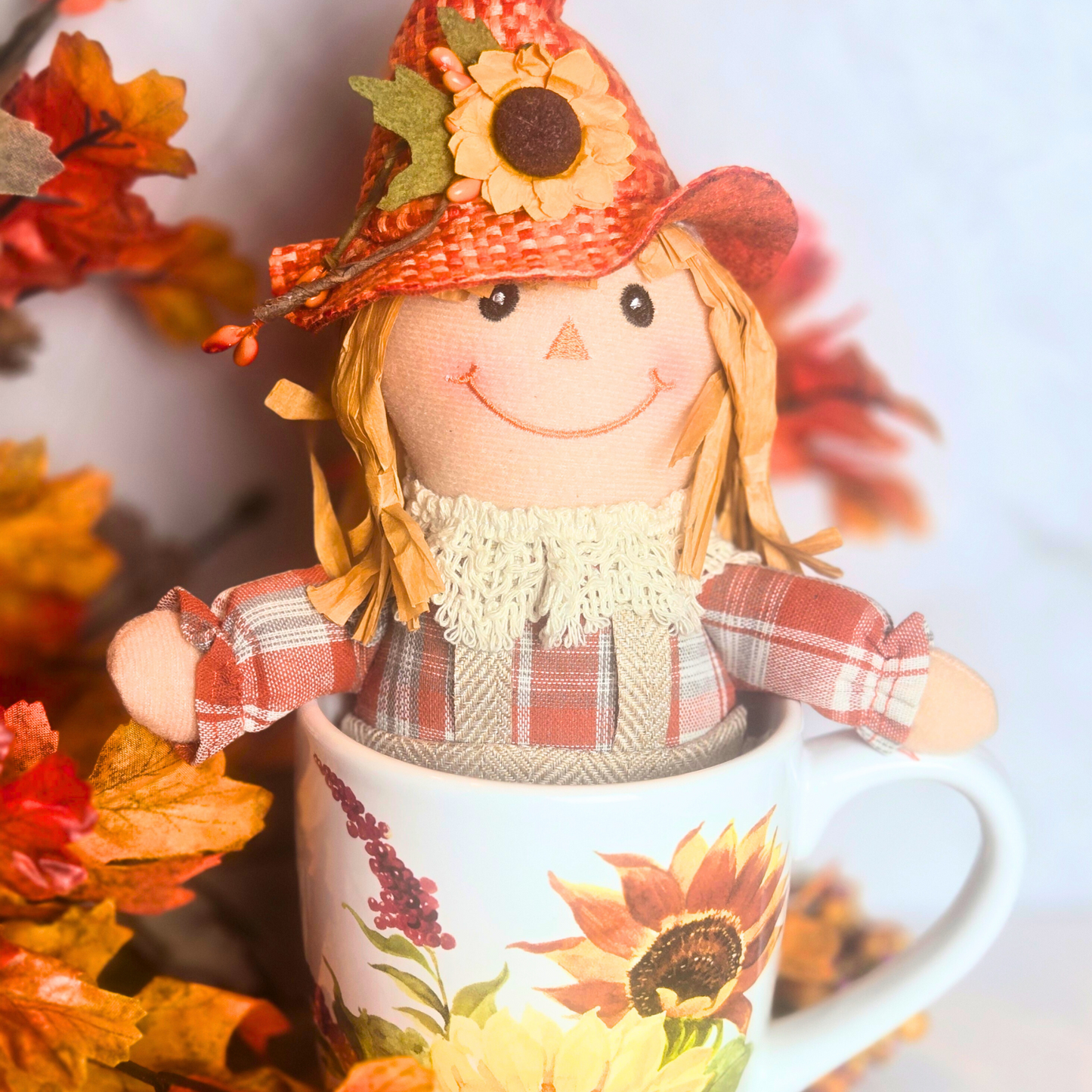 Scarecrow with sunflower hat on a mug with sunflower design, surrounded by autumn leaves.