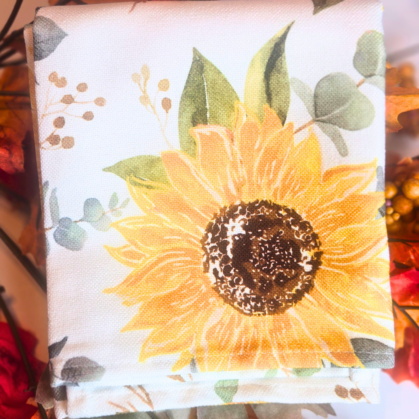 Kitchen towel with a sunflower design on a white background surrounded by fall leaves.