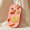 An orange oval-shaped decorative wax sachet with dried flowers and botanical pieces embedded in it, hanging from a white ribbon on a beige fabric background.