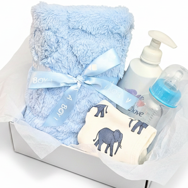 A baby gift set arranged in a box with a fluffy light blue blanket tied with a ribbon,  a folded bib with dark blue elephant prints, and a clear baby bottle with a blue cap.