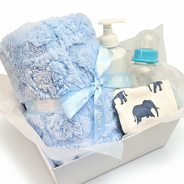 A baby gift basket set containing a light blue plush blanket tied with ribbon, lotion, a clear bottle with a blue cap, and a white bib with navy elephant prints, all arranged in a white box.