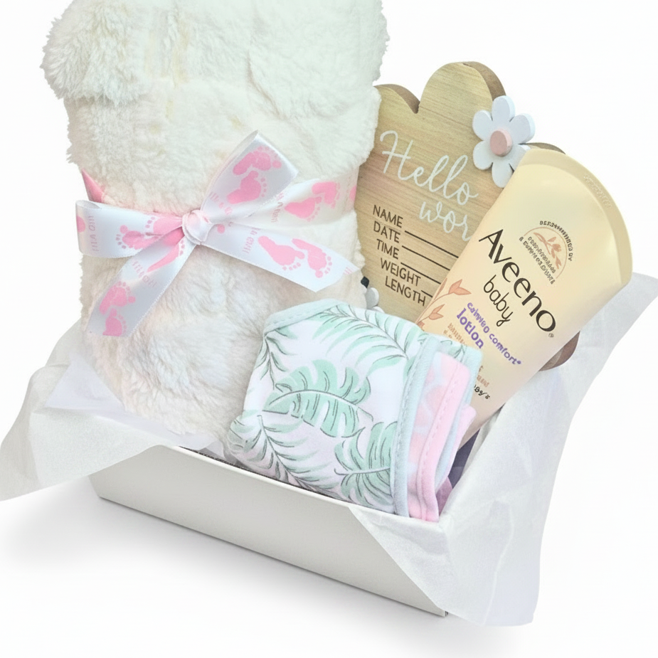 Baby gift basket containing a white blanker with a pink and white ribbon bow, a tube of Aveeno baby lotion, a wooden name/date plaque, a folded leaf-print baby bib, and animal print bib in a white box.
