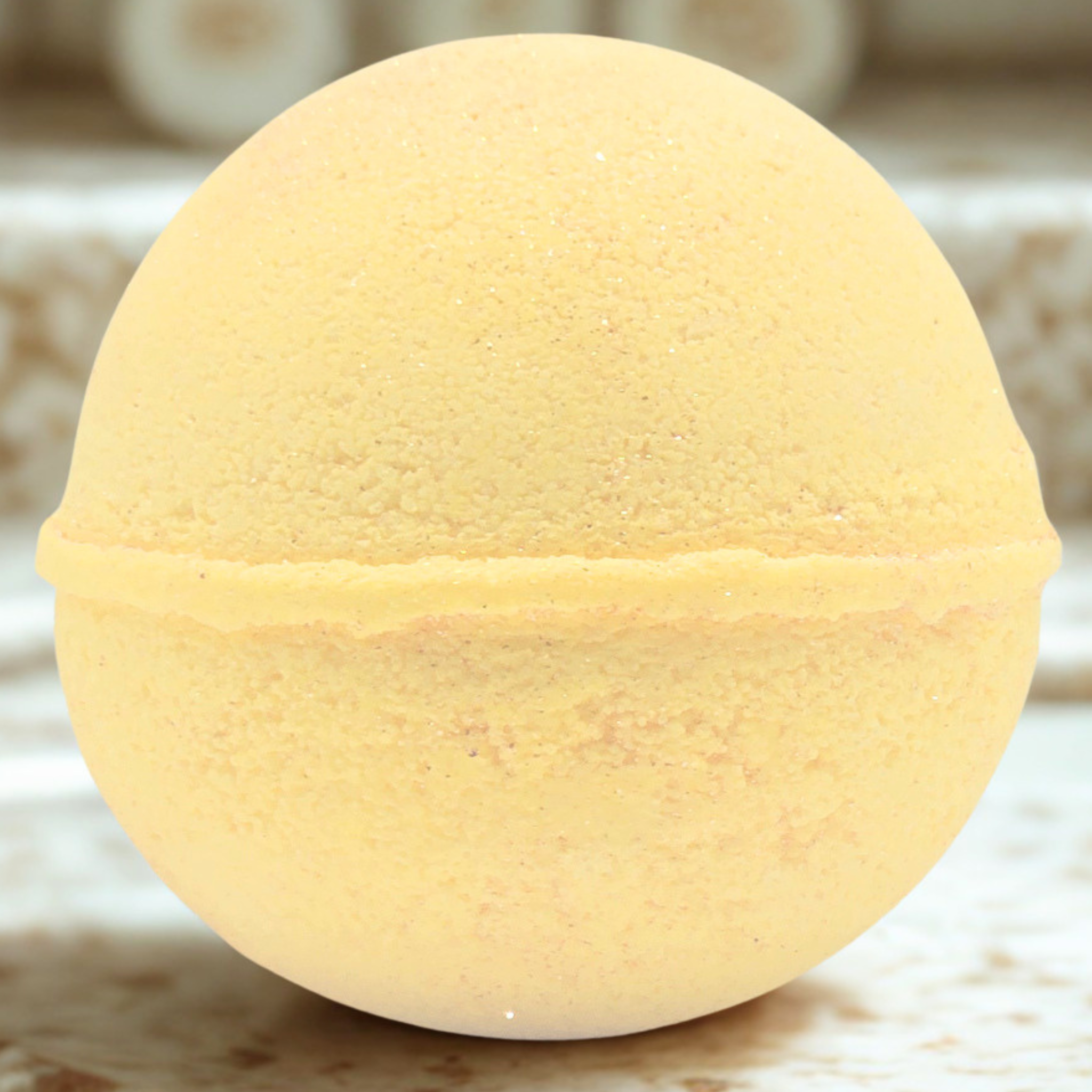 A round, pale yellow bath bomb with a textured, porous surface, resting on a light marble-patterned surface.