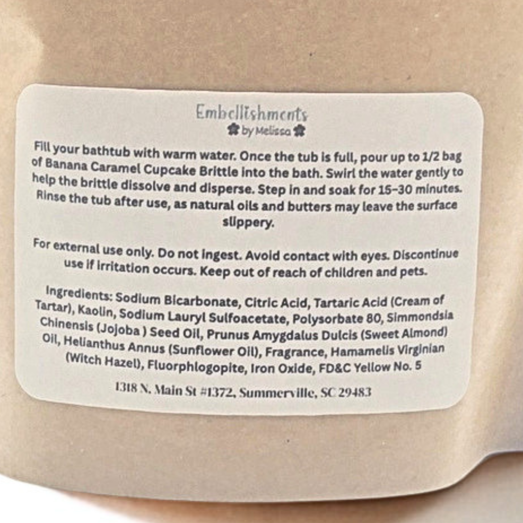 A close-up of a beige paper bag with a white product label reading 'Embellishments by Melissa' and product instructions for banana caramel cupcake brittle.