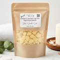 A kraft paper stand-up pouch of Banana Caramel Cupcake Bath Bomb Brittle by Fungal Embellishments, shown on a light marble surface with a white candle, green leaves, and a wooden dish in the background.