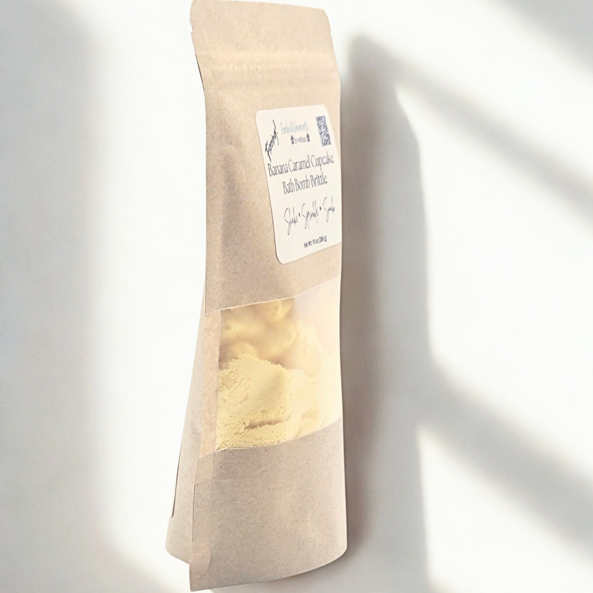 A stand-up kraft paper pouch of bath bomb brittle with a clear window showing yellow pieces inside, photographed against a white wall with soft diagonal shadows.