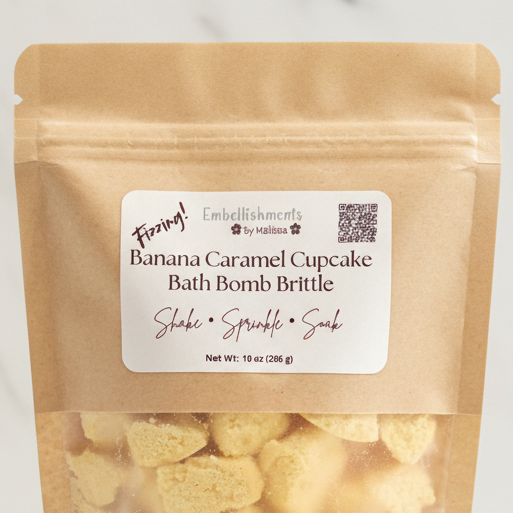 A stand-up kraft paper pouch of Fizzing! by Embellishments by Malina Banana Caramel Cupcake Bath Bomb Brittle, with a white label and visible yellow bath bomb brittle through the clear lower section.