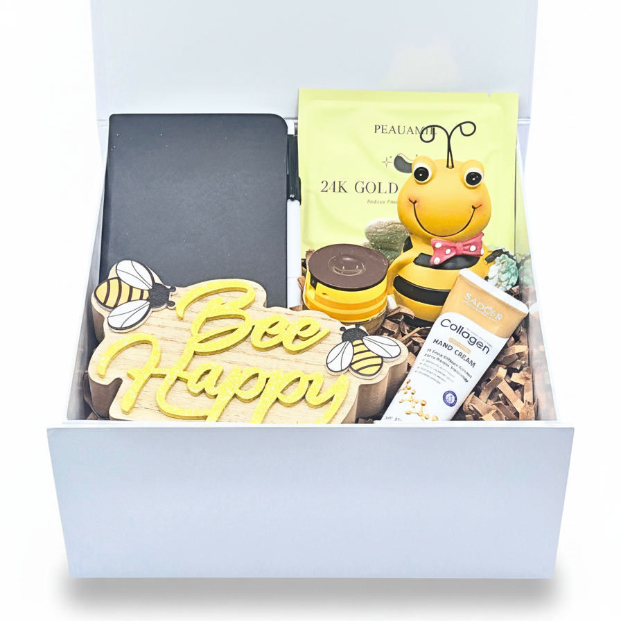 A curated gift box set containing a black notebook, a yellow 'Bee Happy' wooden sign, a '24K GOLD' pouch, bee decor, a tube of hand cream, and lip gloss, all arranged in a white box with shredded brown filler.