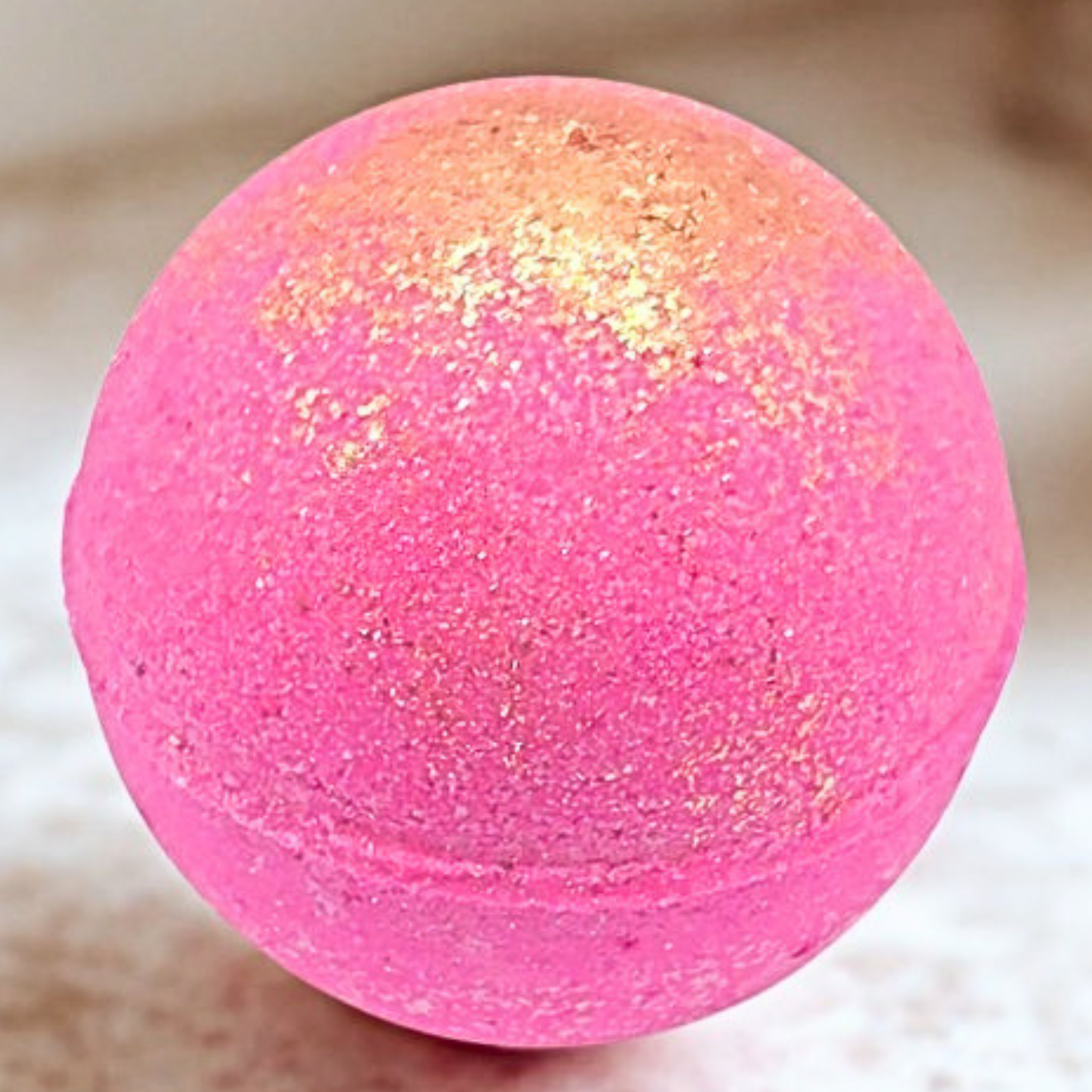 A round pink bath bomb with a textured glitter finish and a gold shimmer concentrated near the top, resting on a light beige surface.