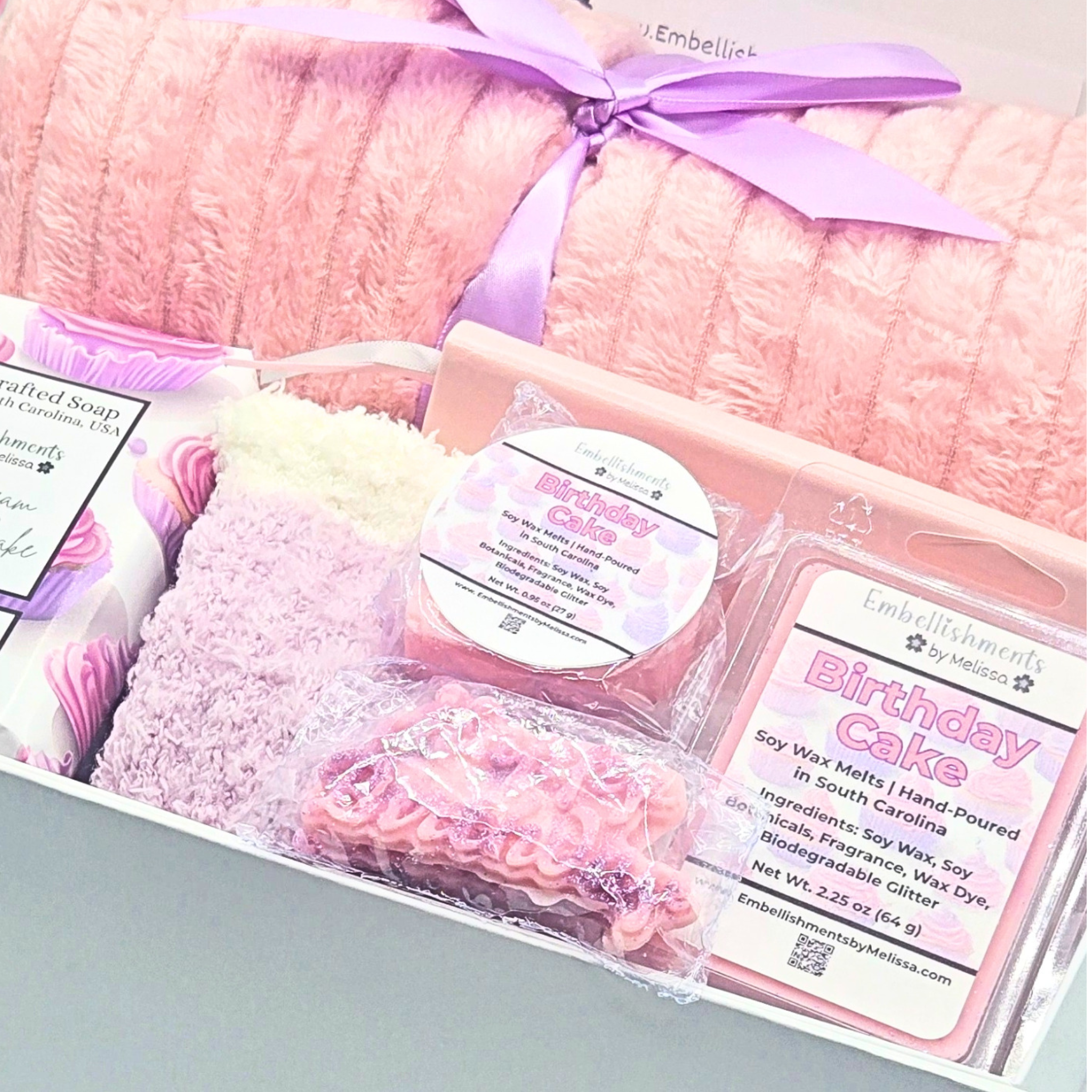 A gift set from Embellishments by Madison featuring a pink plush blanket tied with a purple ribbon, a bar of soap, birthday cake scented wax melt, and a wax melt package inside a white box.