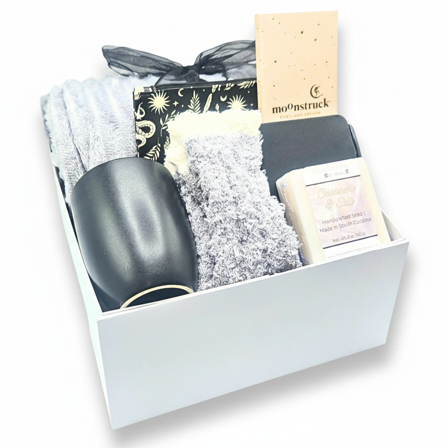 A curated gift box set containing a black mug, cozysocks, a patterned, and a cream-colored Moosinstruck chocolate bar, all arranged inside a white gift box.