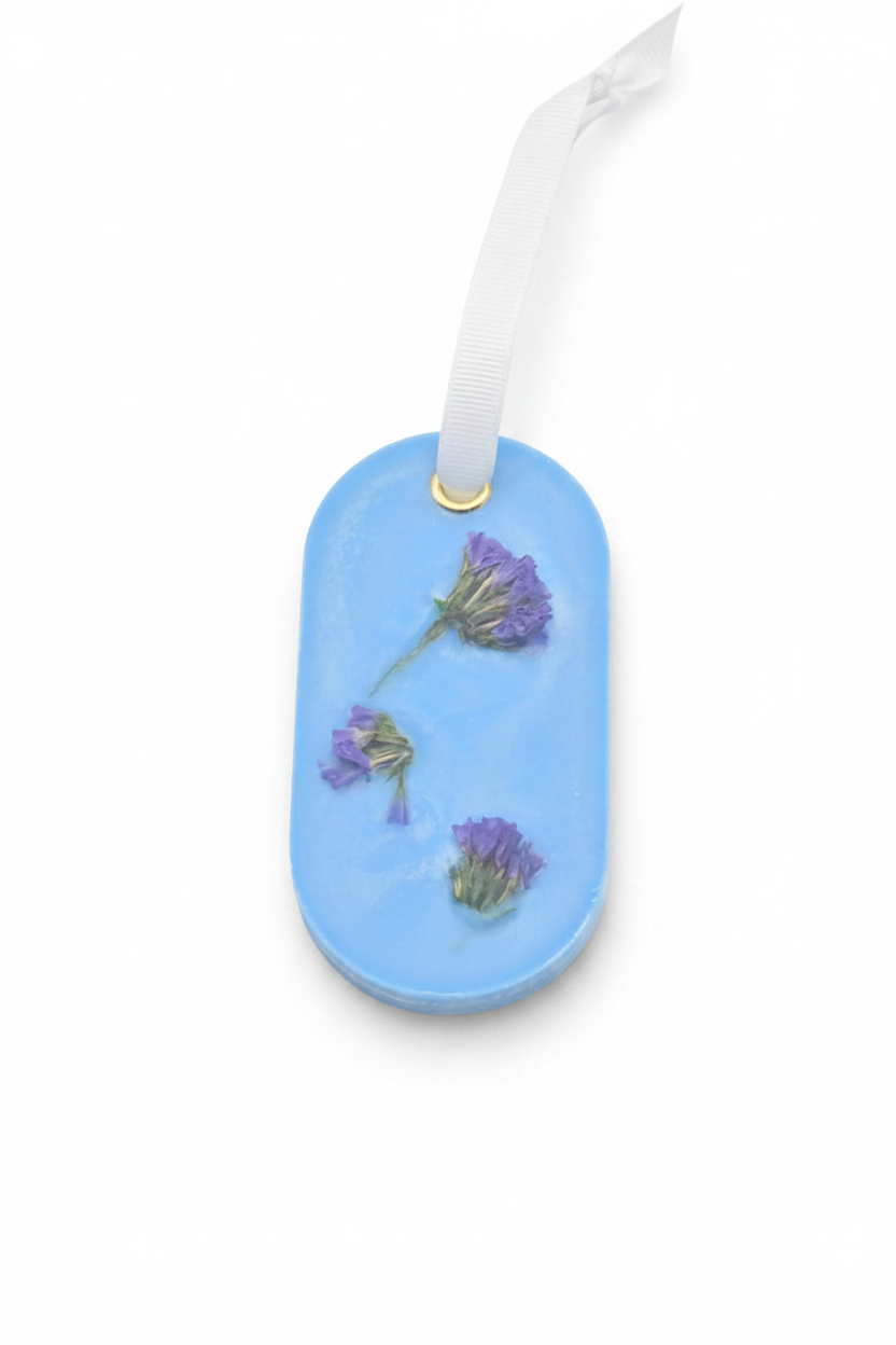 A light blue oval wax sachet featuring three small dried purple flowers embedded, finished with a white ribbon loop at the top.