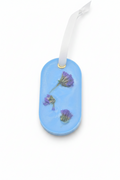 A light blue oval wax sachet featuring three small dried purple flowers embedded, finished with a white ribbon loop at the top.