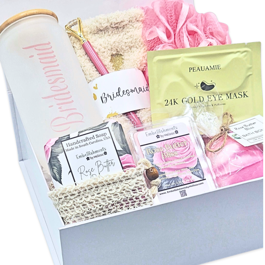 A curated bridal gift set arranged in a white box, featuring a frosted glass tumbler with pink 'Bridesmaid' script, a pink and gold pen, a pink pouf, a gold packet labeled 24K Gold Eye Mask, rose butter soap, and other small gift items.
