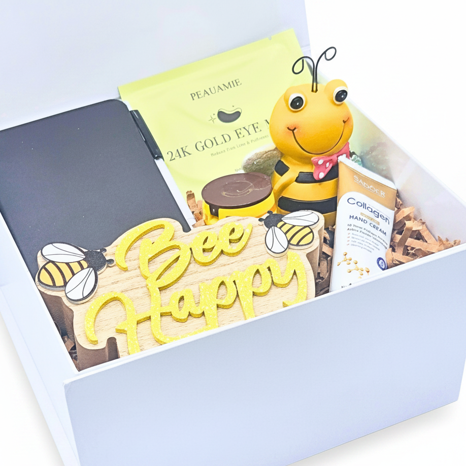 A curated gift box set containing a yellow 'Bee Happy' wooden sign, a 24K gold eye mask packet, a Collagen Hand Cream tube, and a bee figurine, all arranged in a white box with shredded paper.