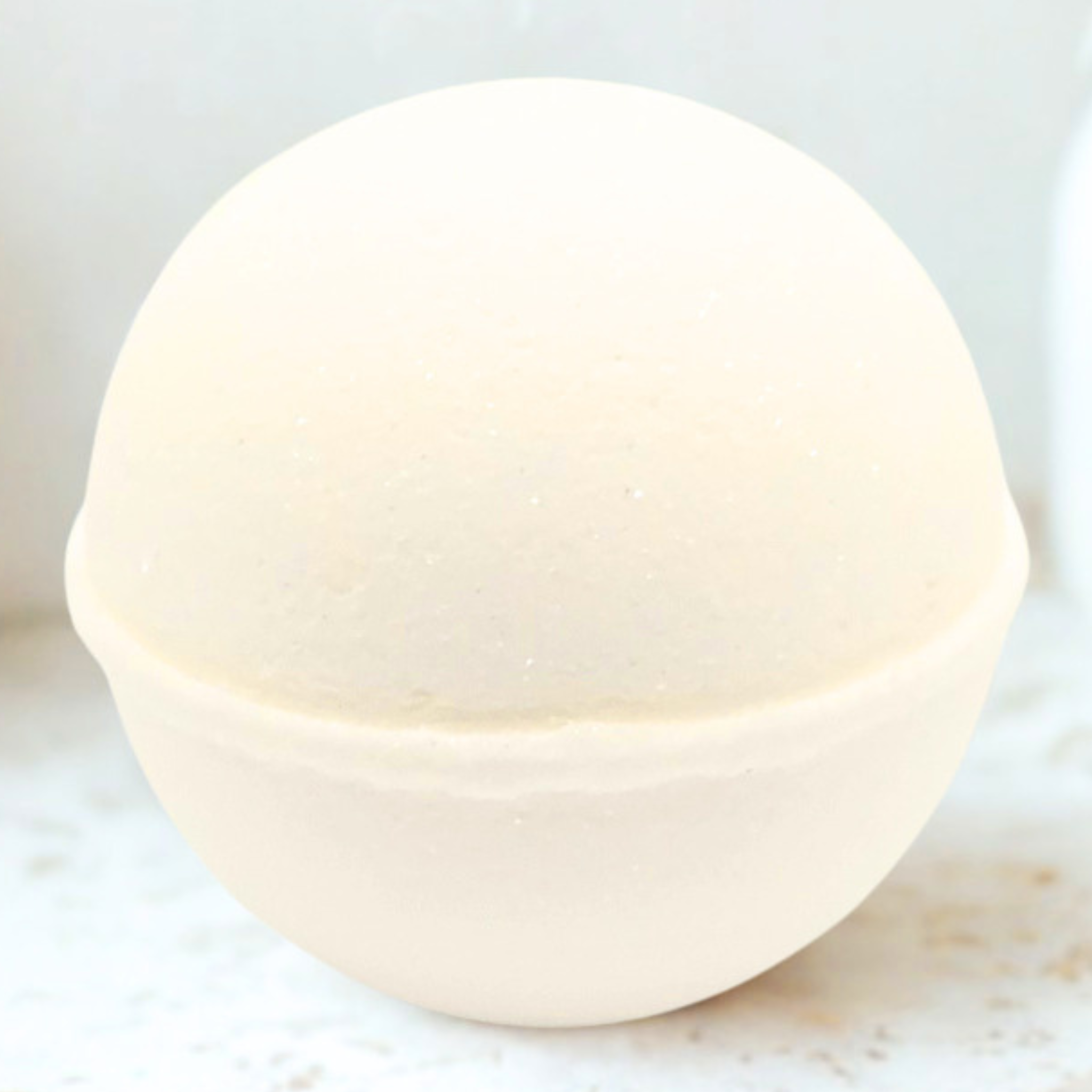A round, off-white bath bomb with a matte, slightly textured surface, resting on a light marble-like surface.