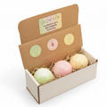 An open kraft gift box containing three pastel bath bombs in a white tray, styled on a beige fabric surface with dried flowers and greenery. The inside lid shows frozen margarita, sangria, and pina colada scents.