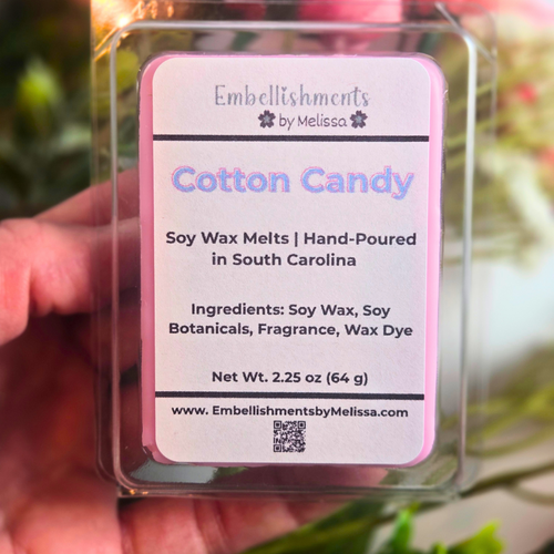 Soy wax melts packaging labeled 'Cotton Candy' by Embellishments by Melissa, held in a hand with a blurred floral background, front view.