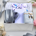 Gift set with a throw, mug, mini notebook with pen, and small affirmation sign on a  grey surface with flowers.