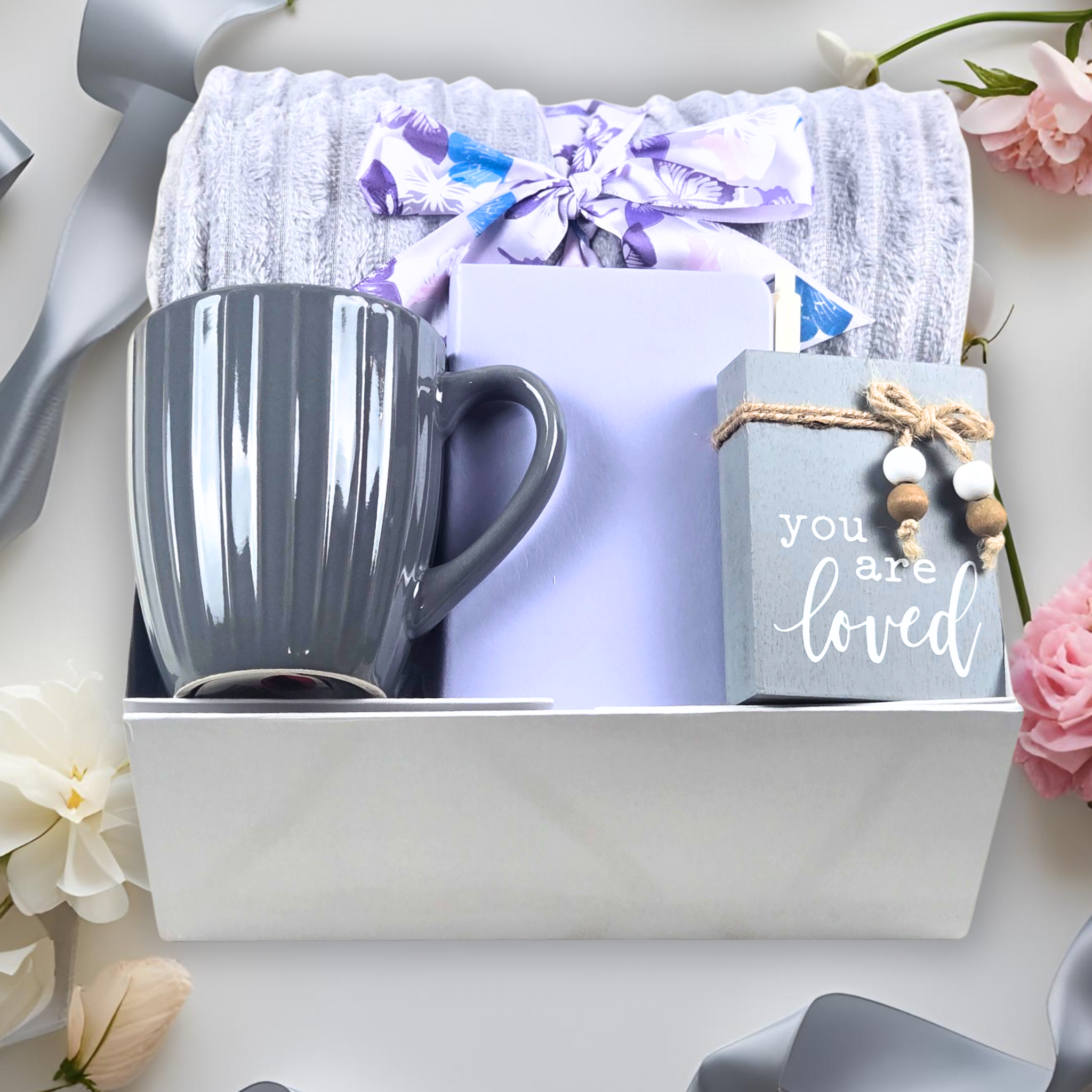 Gift set with a throw, mug, mini notebook with pen, and small affirmation sign on a  grey surface with flowers.