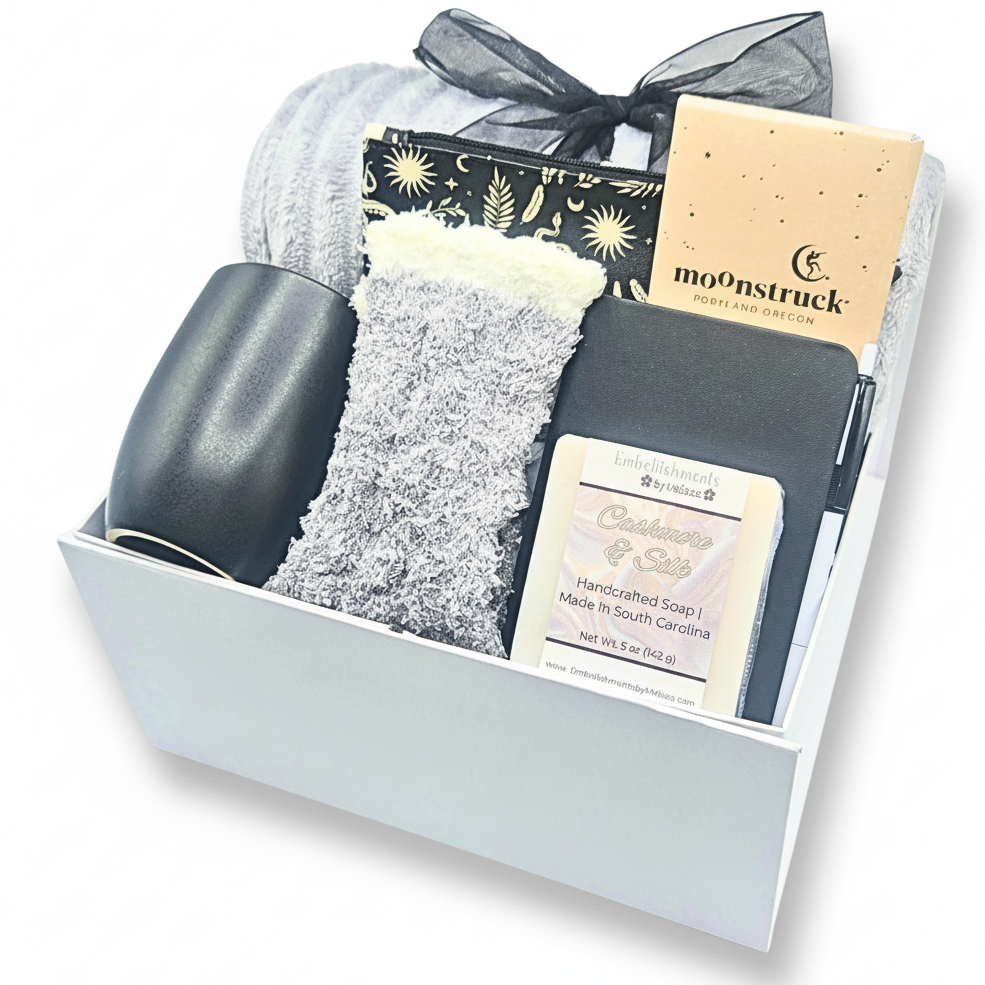 A curated gift box set containing a black mug, cozy socks, a black and gold patterned pouch, a beige chocolate bar labeled moonstruck, and a white bar of soap labeled Handcrafted Soap arranged inside a white box.