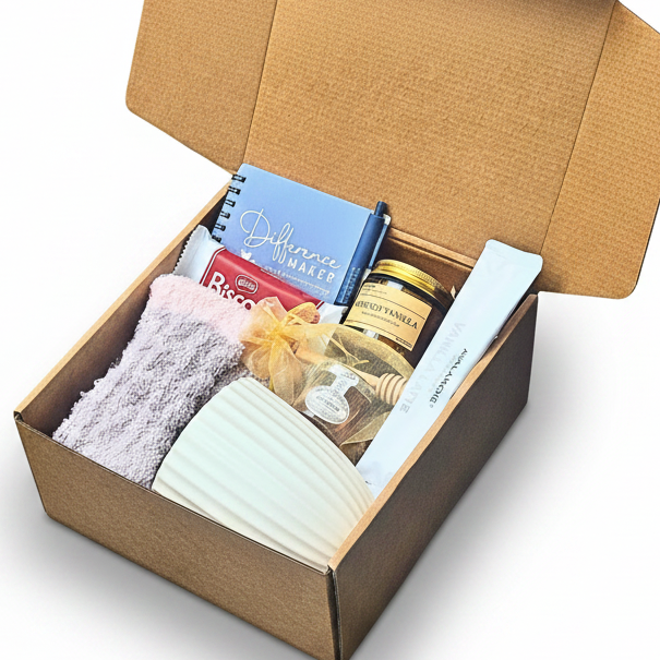 An open brown kraft gift box containing a variety of items, including a blue notebook, french vanilla candle, cozy socks, a white ribbed cup, honey with dipper and Biscoff cookies on a white surface.