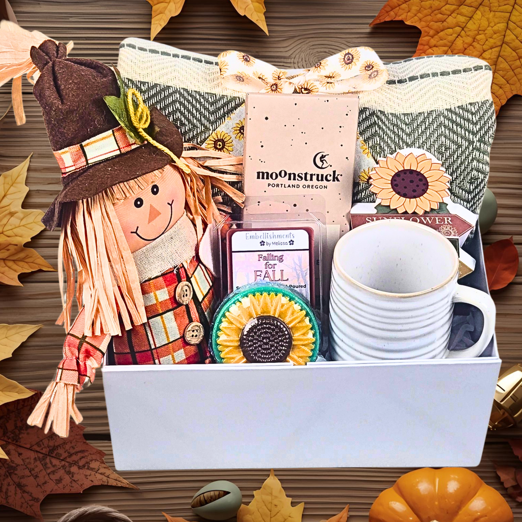 Gift box with fall-themed items including a scarecrow, tea bags, and mugs on a wooden surface with autumn leaves.