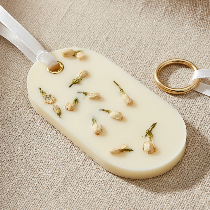 An oval white wax sachet featuring dried flowers embedded inside, suspended from a white ribbon with a gold-colored metal loop at the top, set against a plain white background.