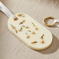 An oval white wax sachet featuring dried flowers embedded inside, suspended from a white ribbon with a gold-colored metal loop at the top, set against a plain white background.