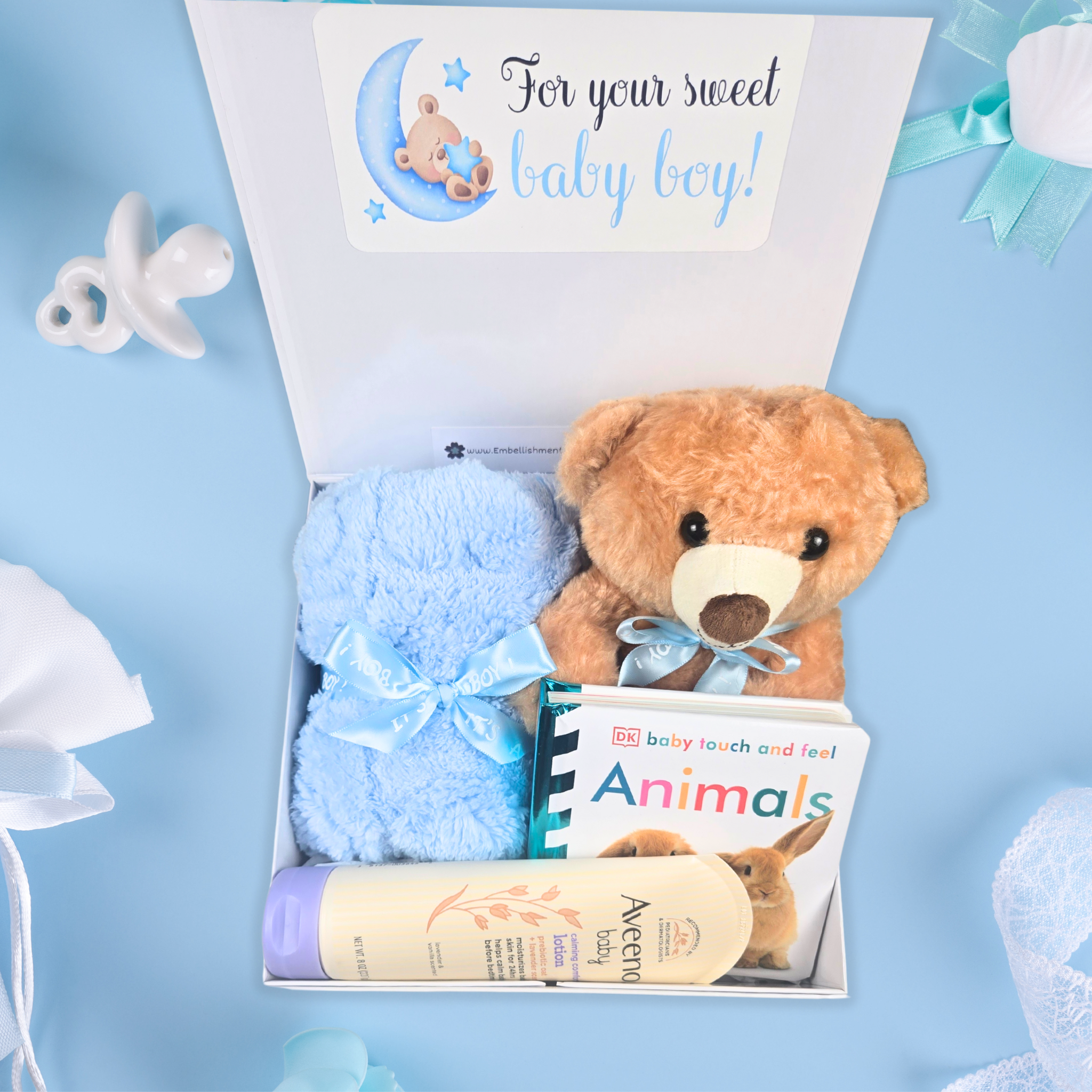 New baby boy essentials box with plush bear, baby blanket, soothing lotion, and book, top view.