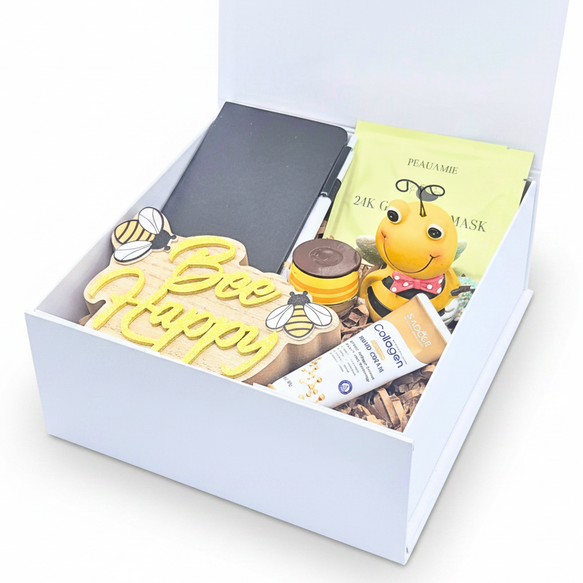 An open white gift box containing a bee-themed gift set including a yellow 'Bee Happy' wooden plaque, and a tube of skin cream, and lip mask, arranged in shredded paper filler.