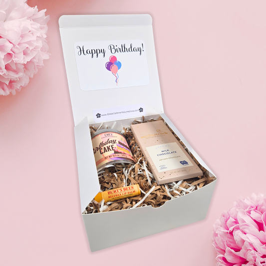 Birthday gift box with large birthday cake scented candle, lip balm and chocolate bar with happy birthday sign on inner lid, side view.