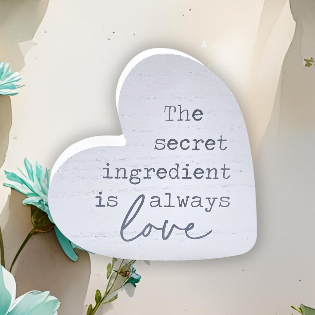 Heart-shaped sign with 'The secret ingredient is always love' text on a light background with flowers.