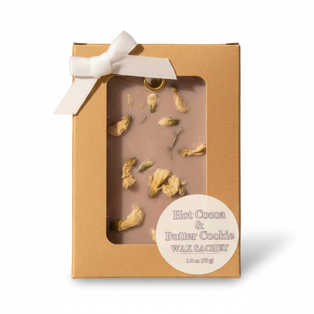 A packaged wax melt product in a kraft paper box with a clear window showing light bown wax sachet eith dried flower petals inside. A white ribbon bow is tied at the top, and a round cream label reads 'Hot Cocoa & Butter Cookie Wax Sachet' on a plain white background.