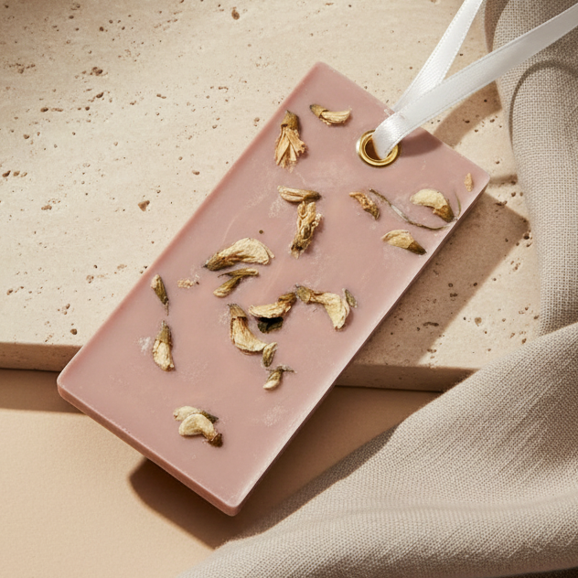 A rectangular brown wax sachet with dried flower petals embedded inside and a white ribbon loop, photographed on a beige stone surface with a beige fabric cloth.