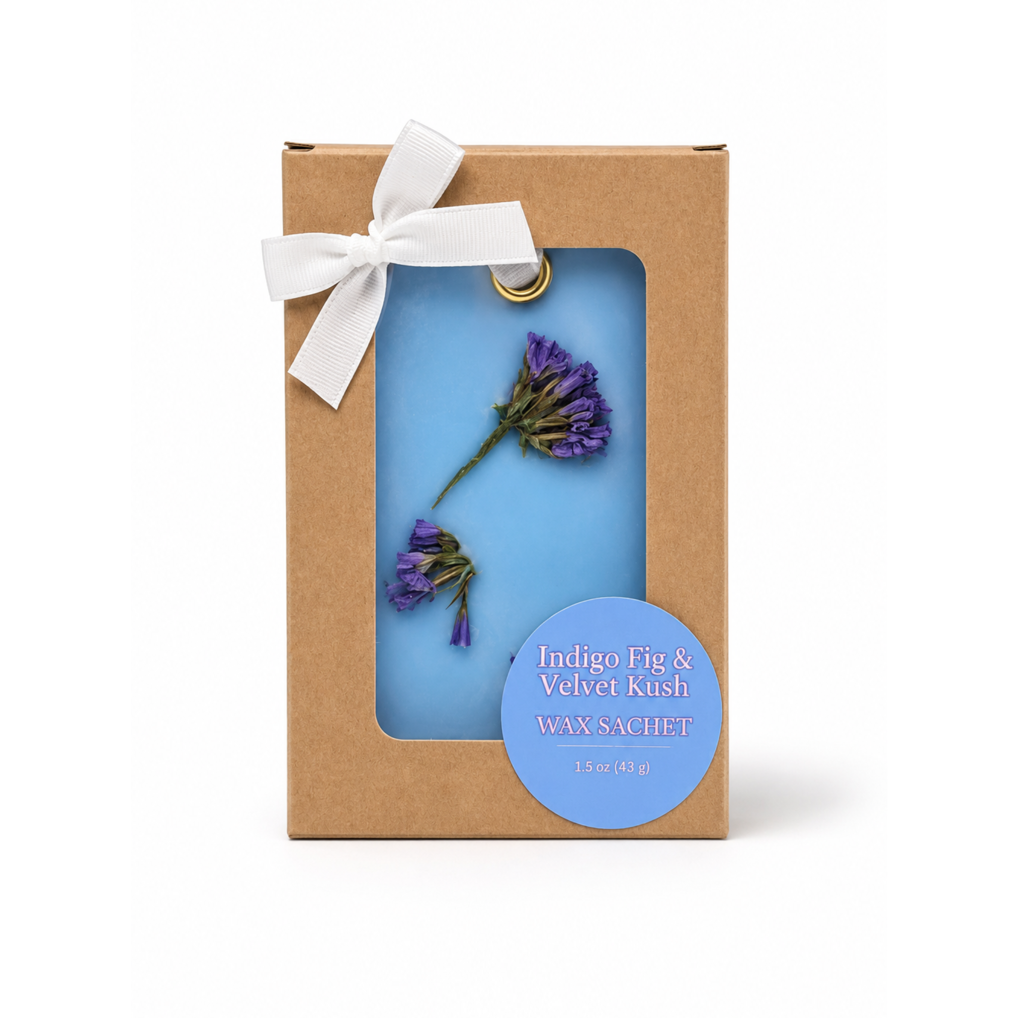 A rectangular kraft paper gift box with a white ribbon bow and a clear window showing a blue wax sachet with purple dried flower buds. A round blue label reads 'Indigo Fig & Velvet Kush WAX SACHET'.