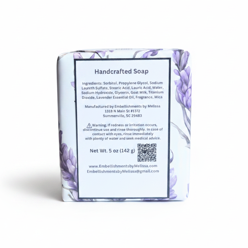 A wrapped bar of handcrafted soap standing upright on a white background. The soap is wrapped in a white paper sleeve with a purple floral pattern and a large white label showing product details, including the brand name 'Embellishments by Melissa'.