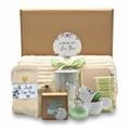 A gift set arranged in front of a large kraft paper box with a floral 'A SPECIAL GIFT For You' sticker. The set includes wax warmer, mint eucalyptus wax melts, a cream-colored blanket with a sage green ribbon, a mug, a soap bar with soap saver bag, a small succulent, cozy socks, a bath bomb, and notebook with pen.