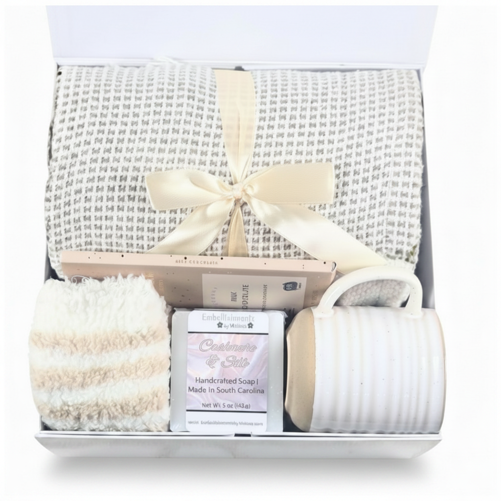 A gift set in a white box featuring a cream patterned blanket tied with a cream ribbon, a striped cream and beige cozy socks, hancrafted soap, a ceramic mug and a chocolate bar.