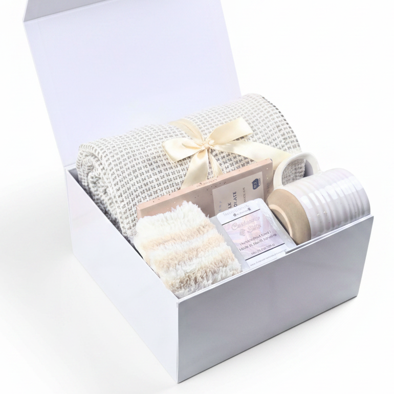 A neutral gift set in a white box, featuring a cream throw tied with a satin ribbon, cozy socks, a mug, handcrafted goat milk soap, and a chocolate bar.
