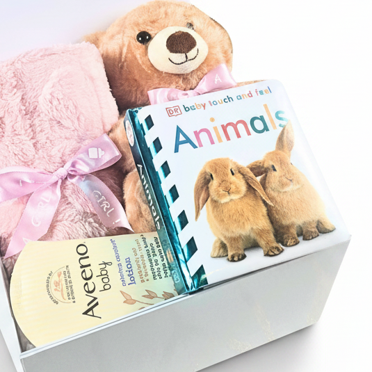 A baby gift basket set in a white box containing a plush brown teddy bear, a soft pink blanket tied with a pink ribbon, a touch and feel Animals children's book, and an Aveeno baby lotion tube.