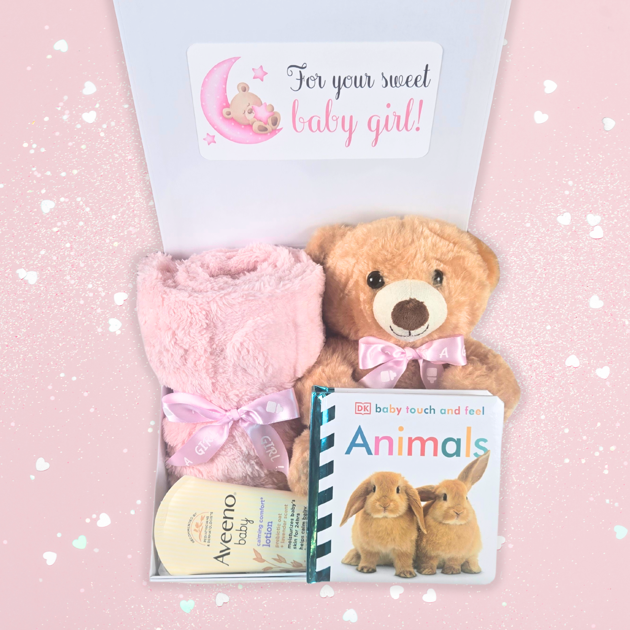 Lovely pink gift box for baby girls, featuring a teddy bear, blanket, soothing lotion, and storybook, top view.