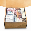 A curated gift box for nurses, shown open with its cardboard lid behind it. The box contains a white mug, a boxed Milk Chocolate bar, a vanilla latte flavored stick, a bracelet, and a blue and white pen, all arranged in brown paper shred.
