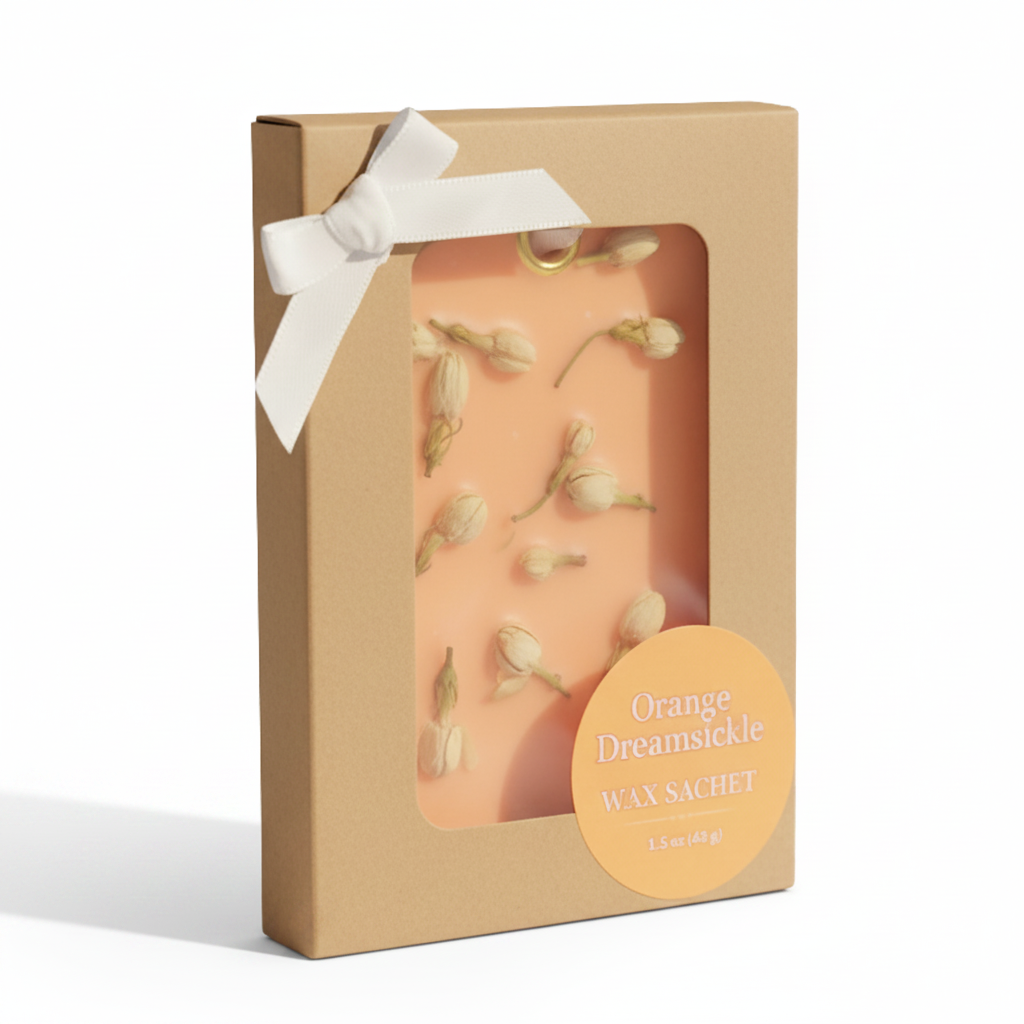A wax sachet in a kraft paper gift box with a white ribbon bow and a clear window showing orange-colored wax with dried flower buds. A round label reads 'Orange Dreamsickle WAX SACHET'.