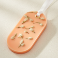 An oval orange-colored wax sachet with dried white flower buds embedded inside, featuring a white ribbon loop at the top.