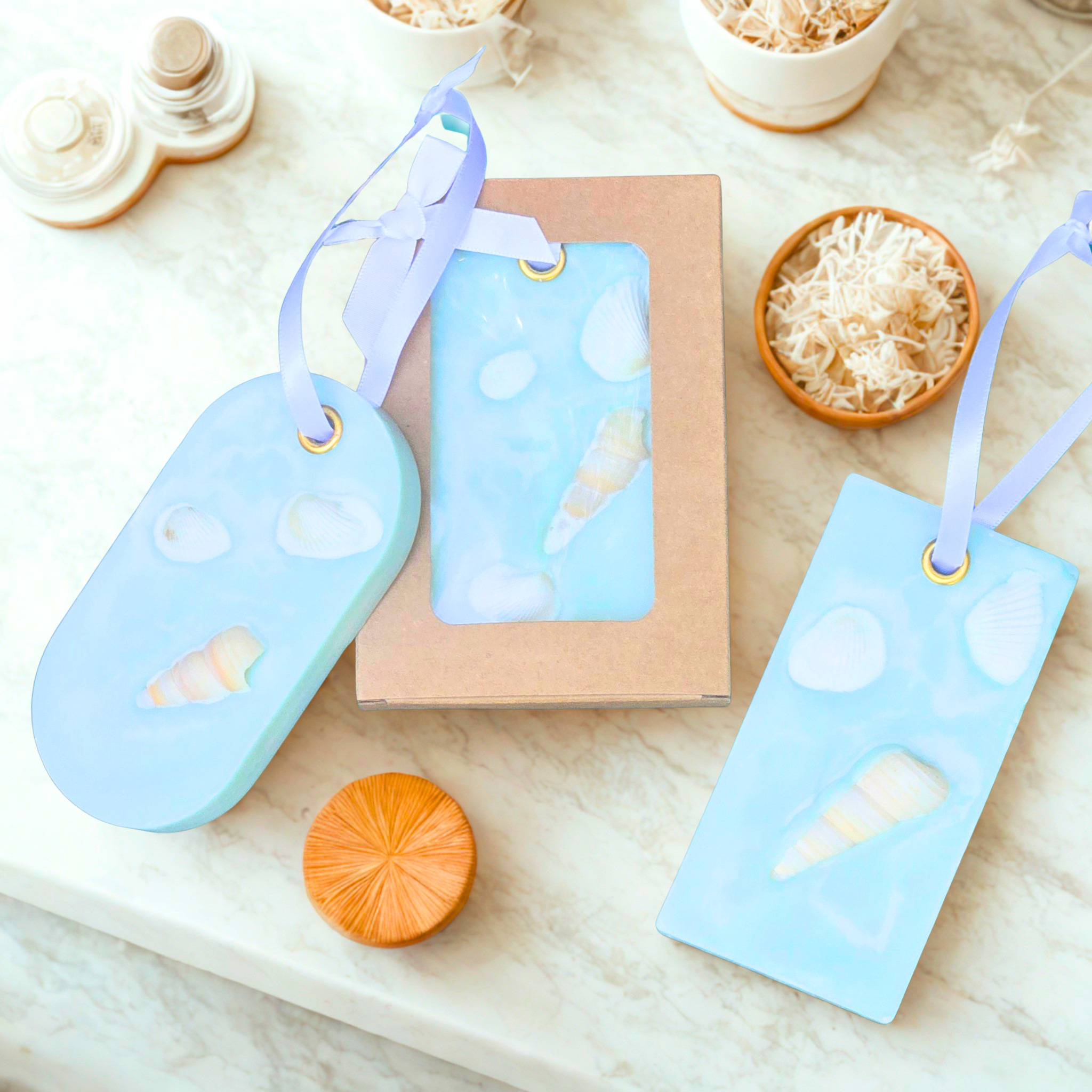 Three pina colada wax sachets with seashell and bubble design with white ribbons, arranged on a light marble surface with small decorative props.