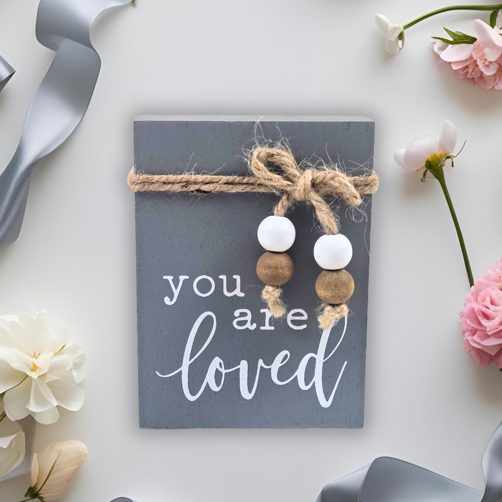 Decorative sign with 'you are loved' text, beads, and a bow on a white background with flowers.