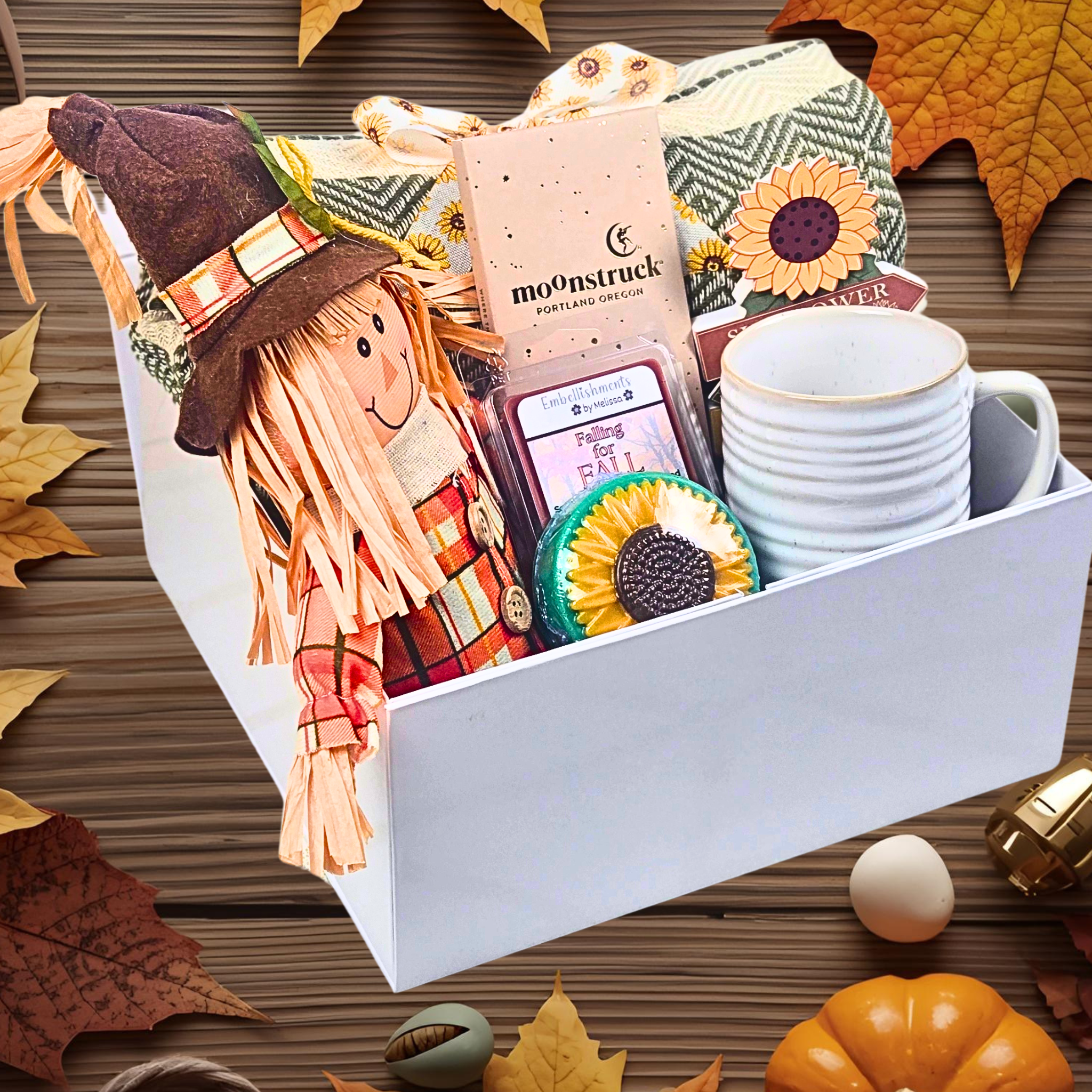 Gift box with a scarecrow, mug, and decorative items on a wooden surface with autumn leaves.