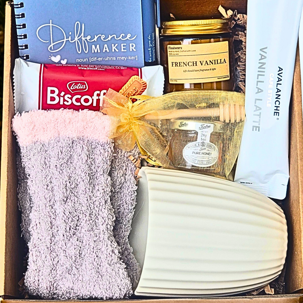 A curated gift box filled with assorted items including a blue Difference Maker notebook, a french vanilla candle, honey with dipper, a pack of Avalanche Vanilla Latte, fuzzy socks, a white ribbed mug and Biscoff cookies.